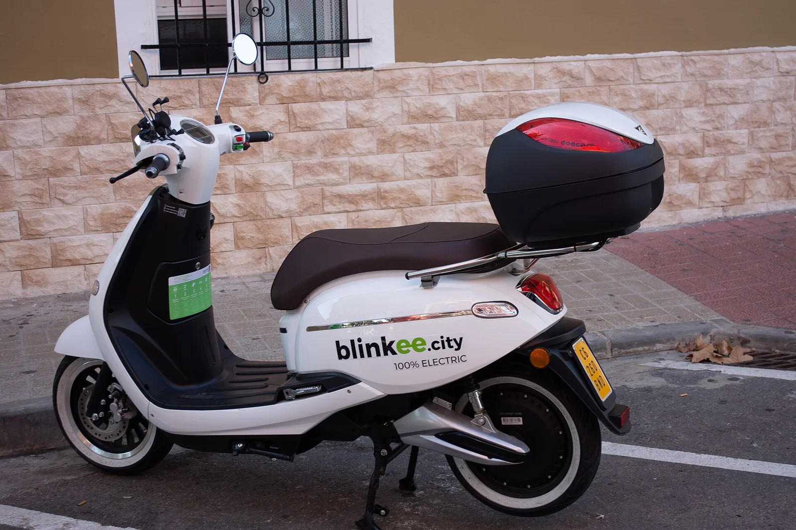Scooter électrique blanc avec top-case garé le long d'un trottoir devant un mur en pierre