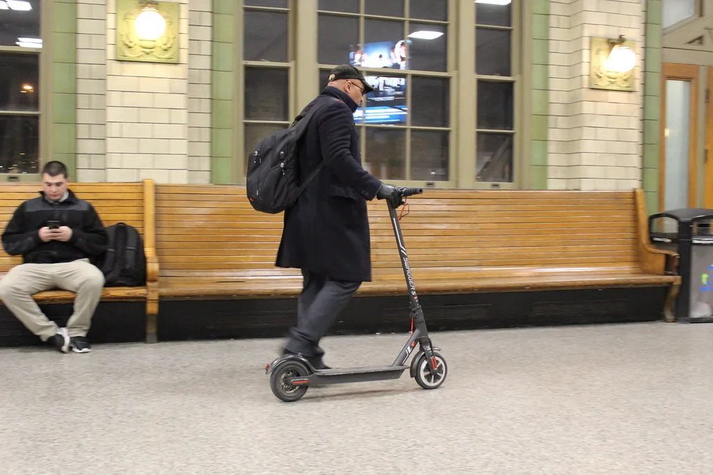 Usager avec sac à dos roulant en trottinette électrique dans le hall d'une gare