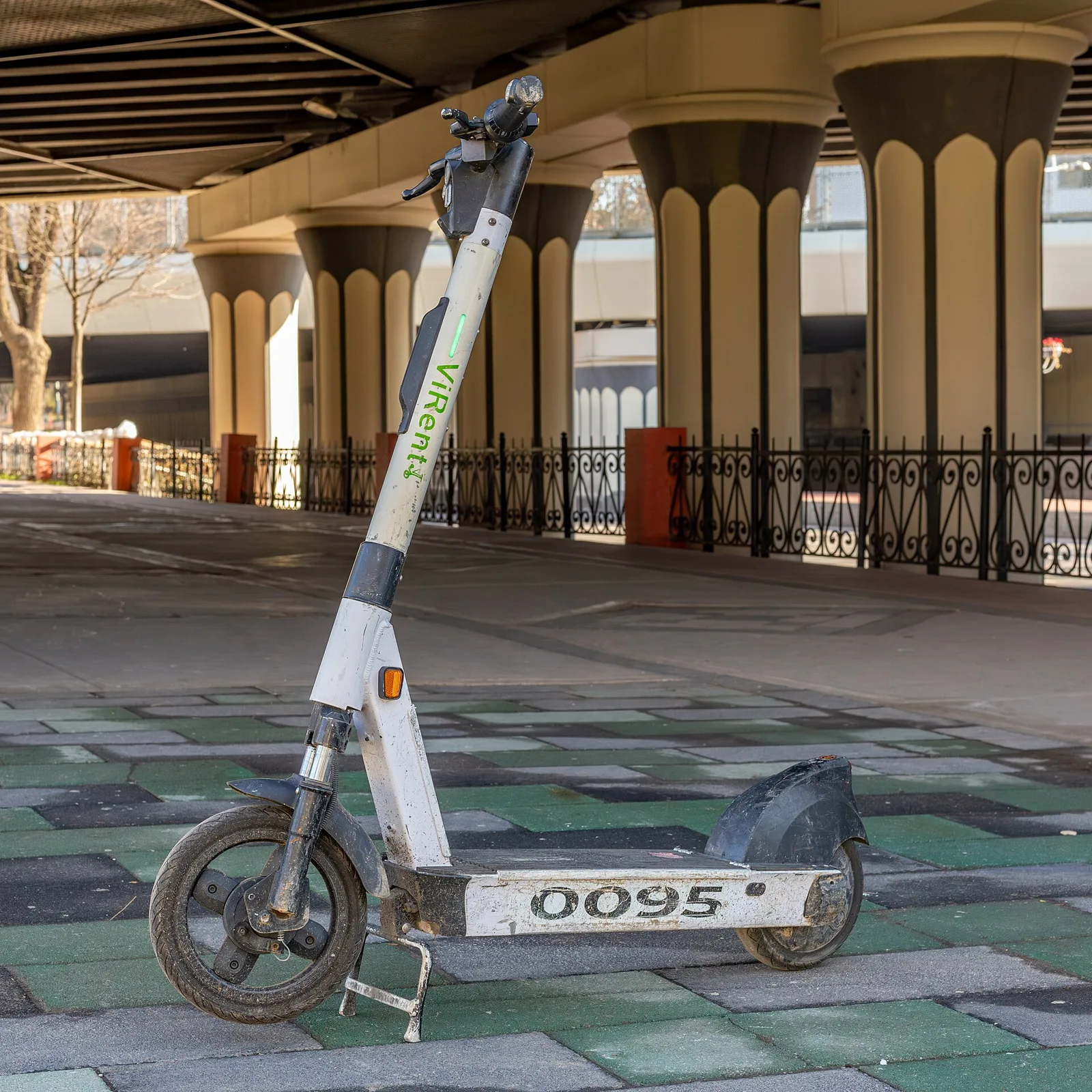 Trottinette électrique blanche garée sur béquille sous un pont, roues et plateau bien visibles