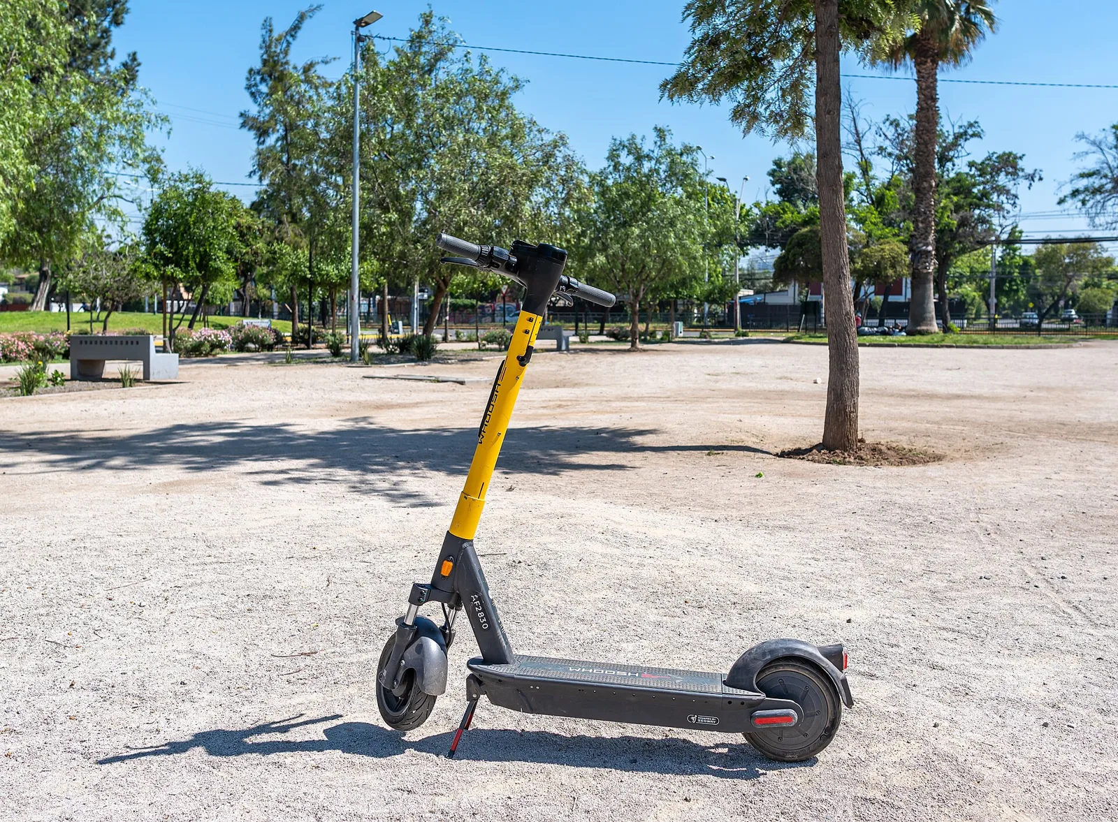 Trottinette électrique grise à tige jaune posée sur sa béquille dans un parc ensoleillé