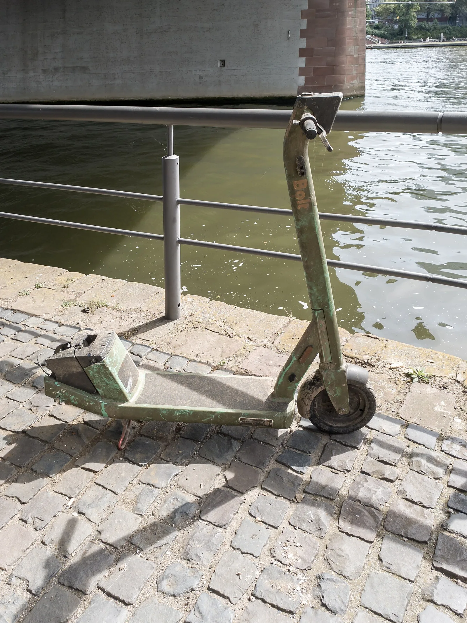 Trottinette électrique usée appuyée contre une rambarde au bord de l'eau sur des pavés