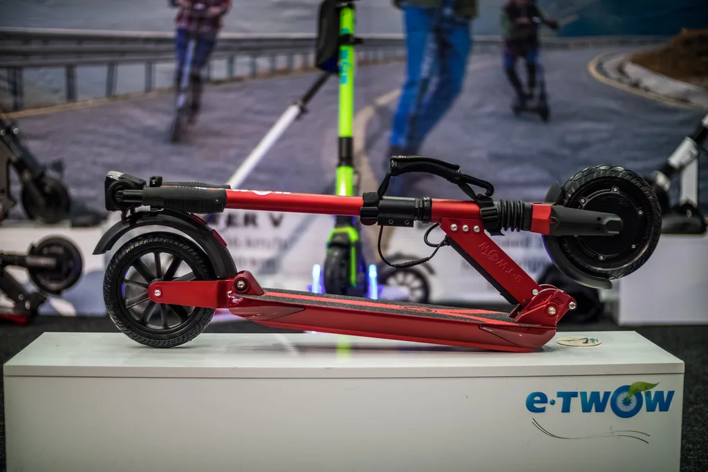 Trottinette électrique rouge pliée posée sur un socle lors d'un salon, vue de profil